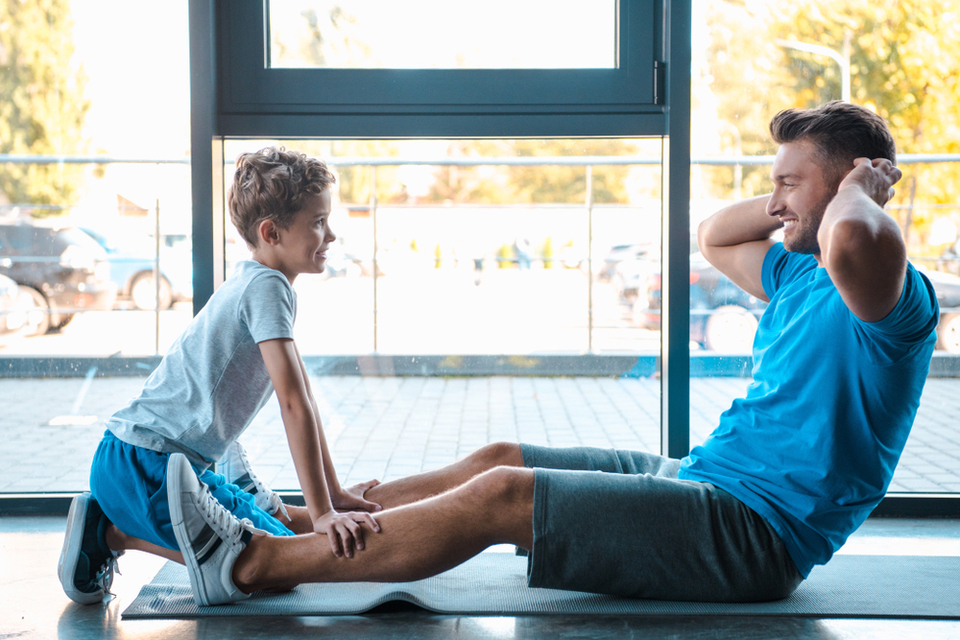 Boy holding down his dad's legs as the dad does sit ups