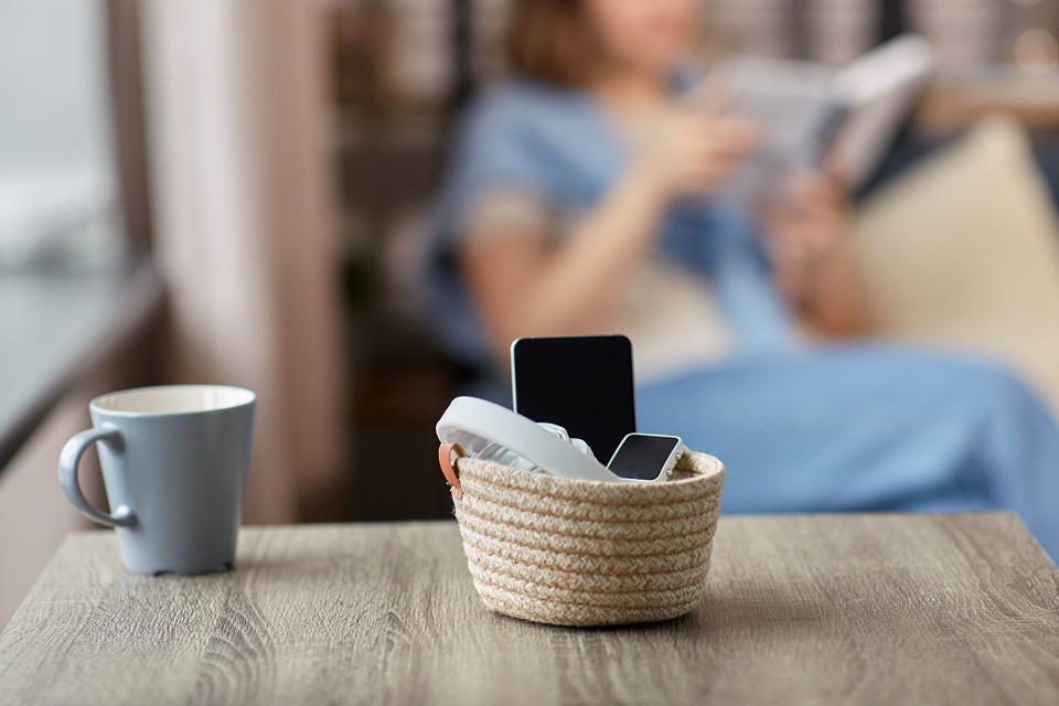 Social Media detox concept: Close up of gadgets in basket on table and woman reading book at home